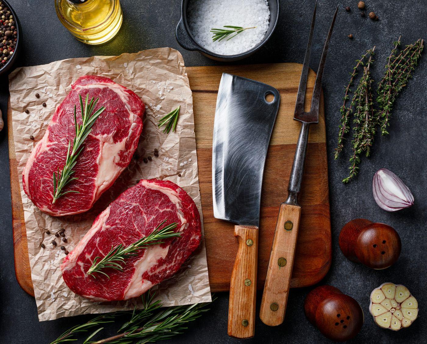 Two rib eye steaks on butcher&rsquo;s paper beside a cleaver and a carving fork